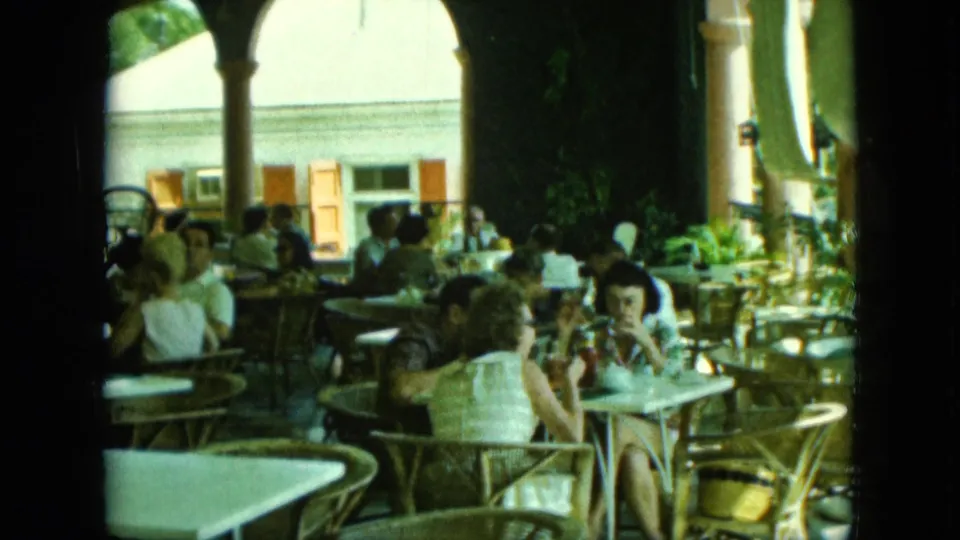 1960s 8mm frame of a shaded outdoor café where patrons sit at wicker tables beneath arched columns.