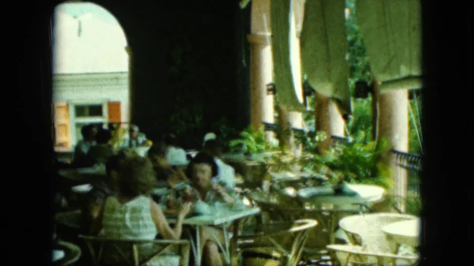 A handheld 8mm view looks across occupied café tables, shaded columns, plants, and a bright building facade.
