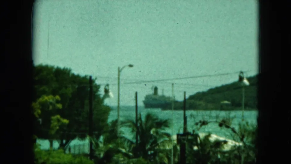 1964 8mm harbor view identified by metadata as Saint Thomas, framed by dark bars with foliage and a distant ship across the water.