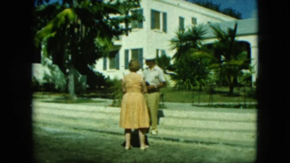 A woman in a yellow dress and a man in light clothing appear to talk outside a white building with palms and lawn.