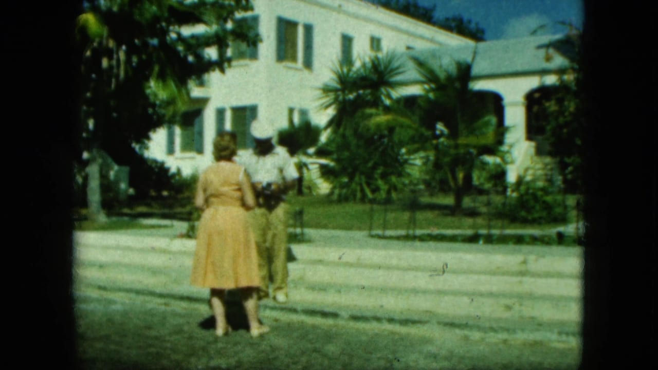 Two adults face each other outside a white building with palms.