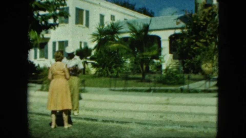 The adults are partly shifted in frame as the camera holds on the white building, greenery, curb, and sunny exterior setting.