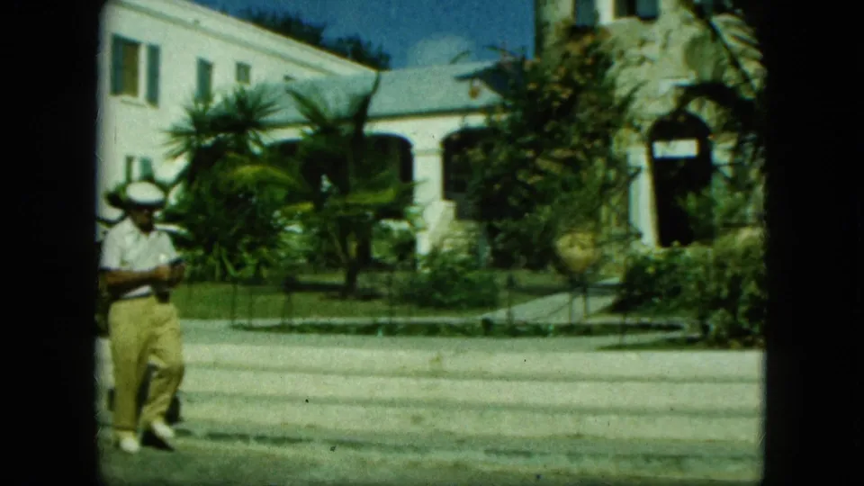 The man appears to step or walk near the curb as the white building, lawn, palms, and stone-edged entry remain visible.