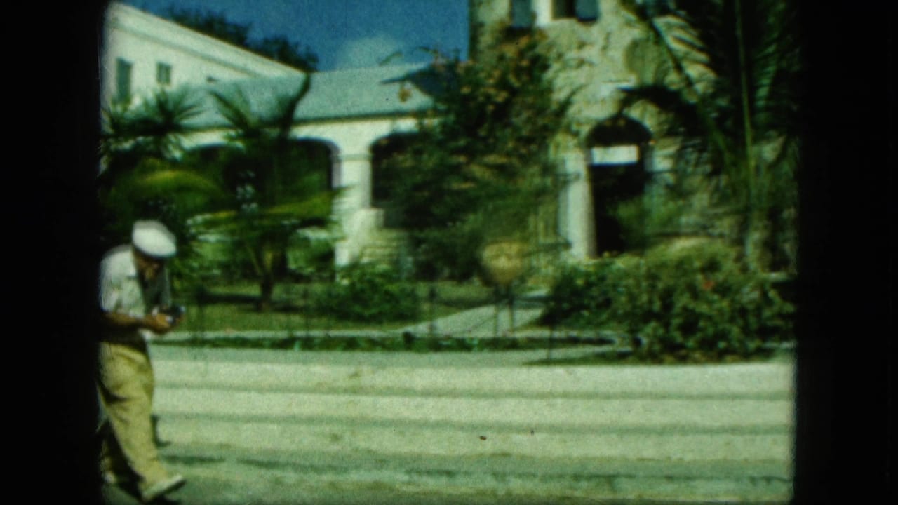 Man in a white cap walks past palms and a stone tower near a white arched building.