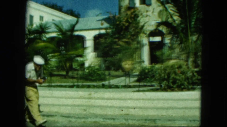 1964 8mm frame from Saint Thomas showing a man walking near palms, a stone tower, and a white building with arched openings.