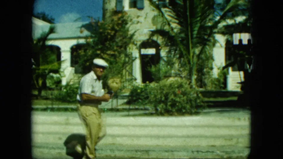 1964 Saint Thomas home-movie frame with a man in a cap crossing a sunlit courtyard before palms, shrubs, and arched stone architecture.
