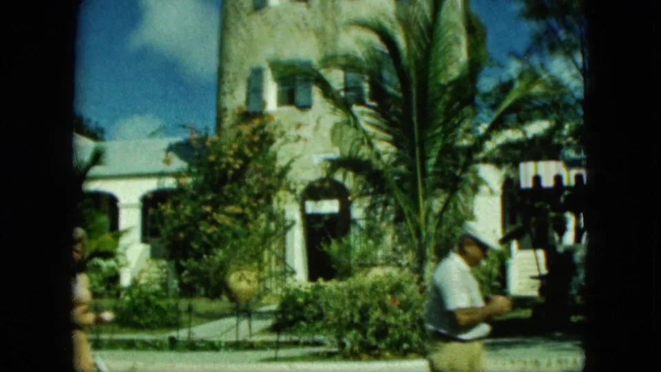 1964 8mm Saint Thomas frame showing the tower and tropical building while a man sits partly in view near the steps.