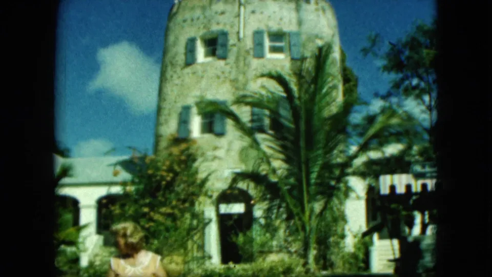 1964 Saint Thomas frame with the stone tower, palms, white building, arched entrance, and a partially visible person in the foreground.
