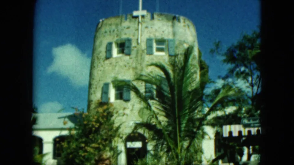 1964 Saint Thomas home-movie frame showing a round stone tower with shuttered windows, roof flags, palm fronds, and bright sky.