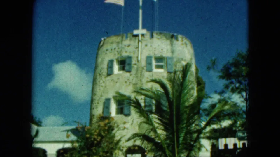 1964 Saint Thomas frame showing the stone tower closer in view, with a cross-like roof detail, windows, palms, and tropical foliage.