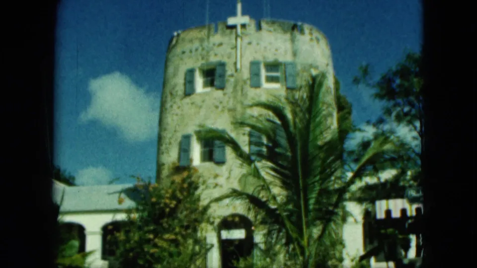Saint Thomas 1964 8mm frame of a historic-looking stone tower with roof flags, palm fronds in front, and trees at the side.