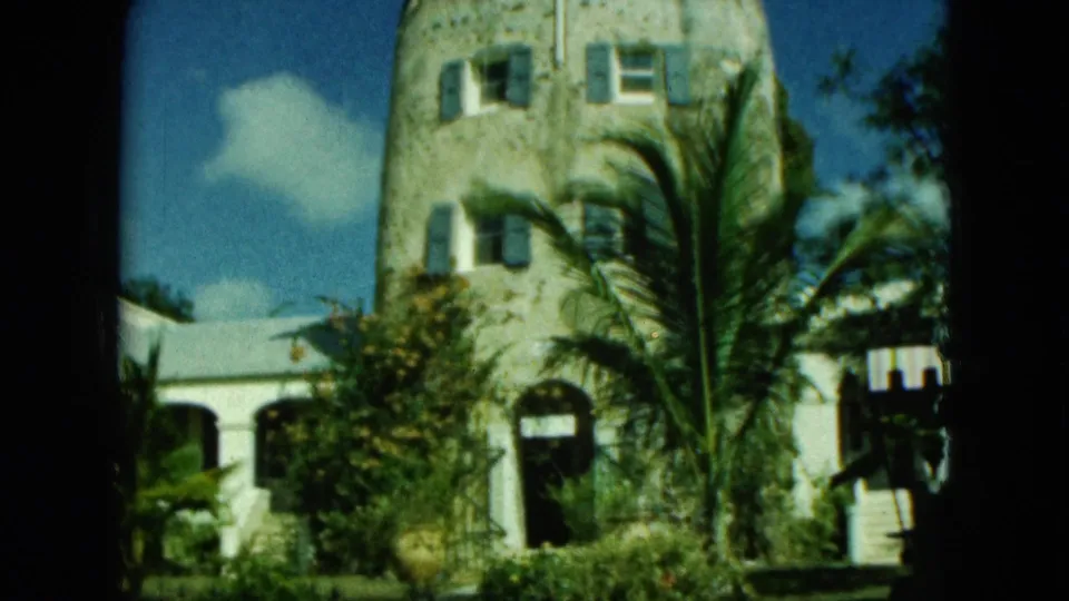 1960s 8mm frame of a tropical stone building facade with shutters, arched entrance, palm trees, and a bright sky.