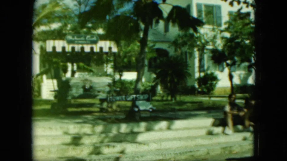 1960s 8mm frame with Tower Gift Shop sign, palms, white building facade, road, small cart, and people seated at the right edge.