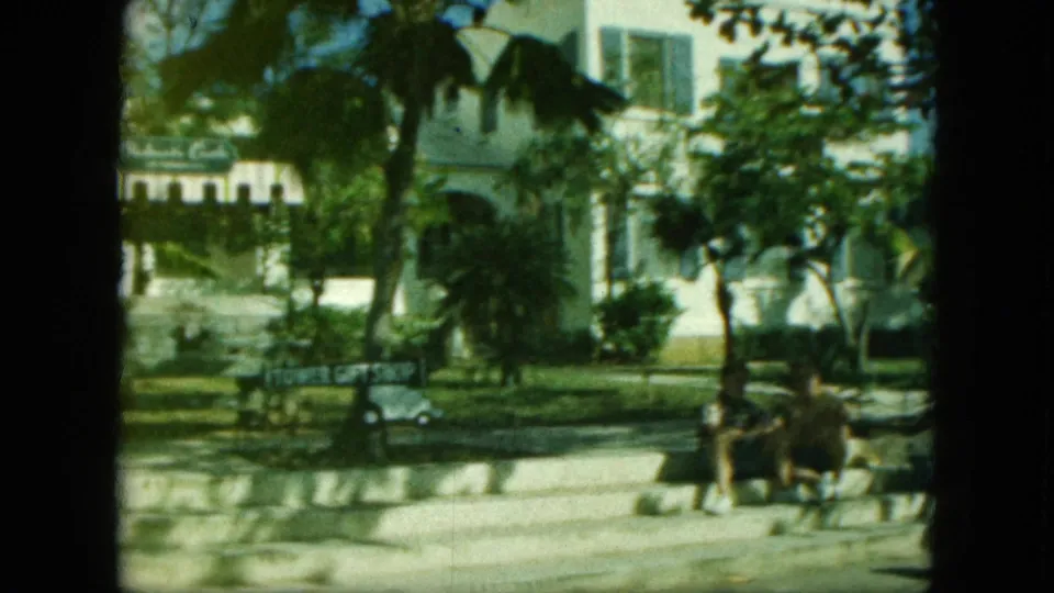 Final 8mm frame showing people seated near the curb, with palms, a white building, street, cart, and Tower Gift Shop sign visible.