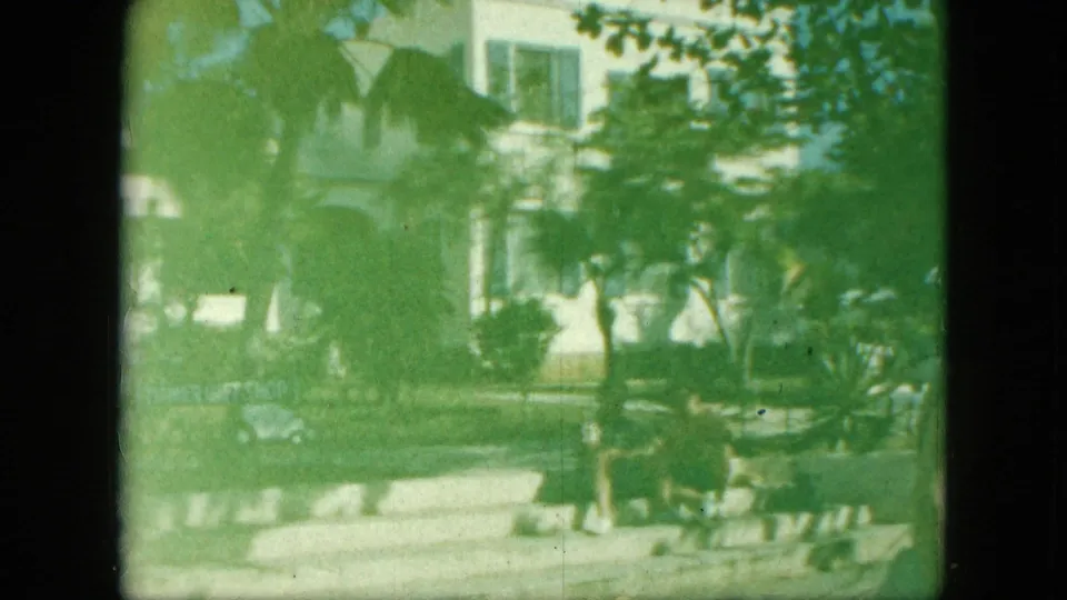 1964 8mm vacation frame with a white building partly obscured by palms and a green exposure shift, with adults seated near steps.