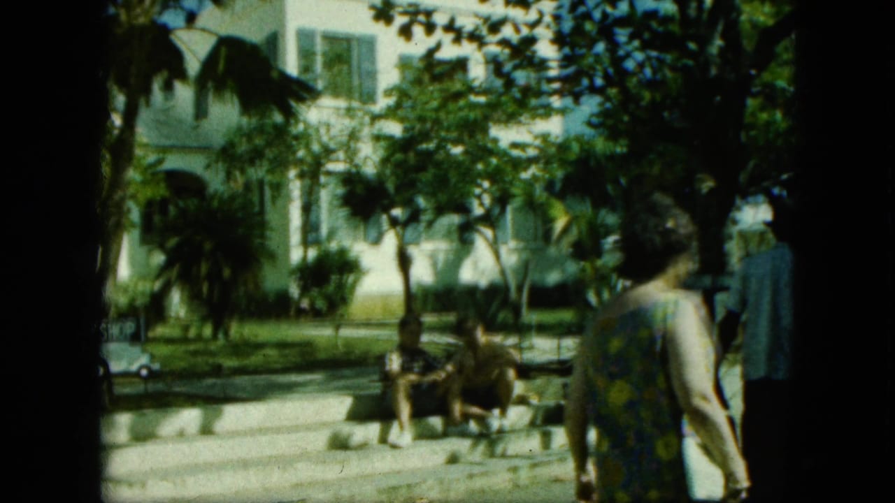Saint Thomas-labeled 1964 home-movie frame showing a woman from behind as she passes seated adults outside a white building with palms.