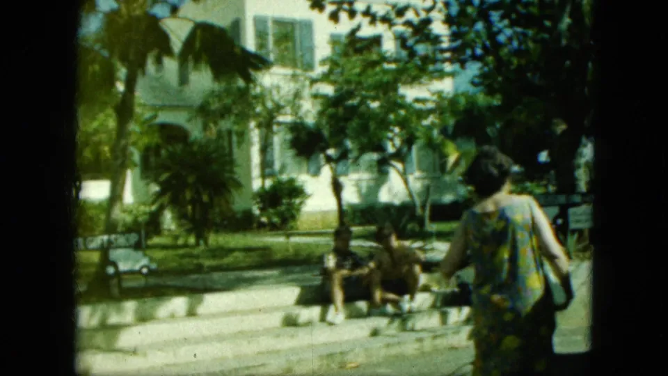 1964 8mm frame from a Saint Thomas-labeled vacation clip, with a woman walking past seated adults near steps, palms, and greenery.