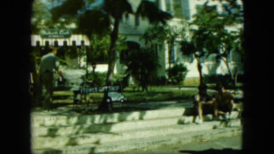 Saint Thomas-labeled 1964 vacation frame with seated adults, palms, a white building, and a readable Tower Gift Shop sign.