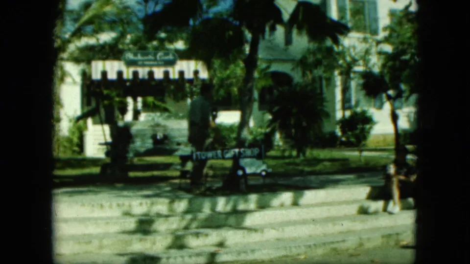 1964 8mm Saint Thomas street view near a tower and Tower Gift Shop sign, with palms, a parked car, and a person walking through the frame.