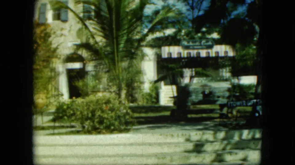 Wider 1964 Saint Thomas view with stone architecture, palms, shrubs, a sidewalk, the Tower Gift Shop sign, and a parked car at the edge.
