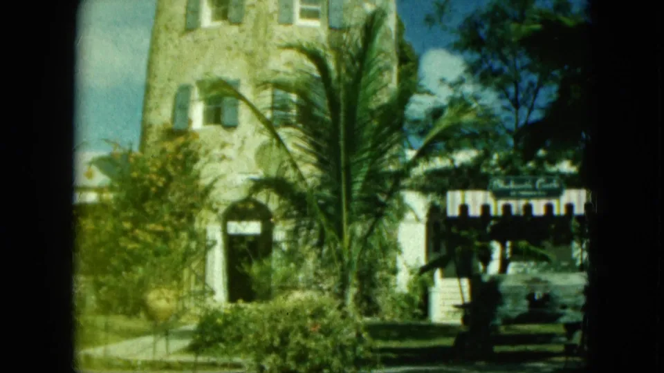 1964 8mm frame near Blackbeard's Castle area in Saint Thomas, showing a stone tower facade, palms, shrubs, and nearby tourist signage.