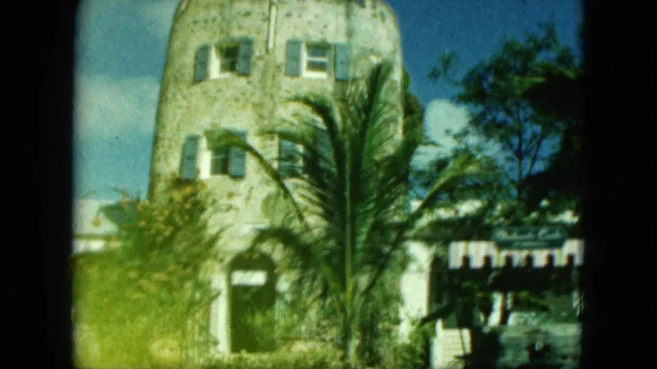 1964 8mm frame of a stone exterior with blue shutters, palm fronds, and a small shaded entry area in Saint Thomas metadata context.
