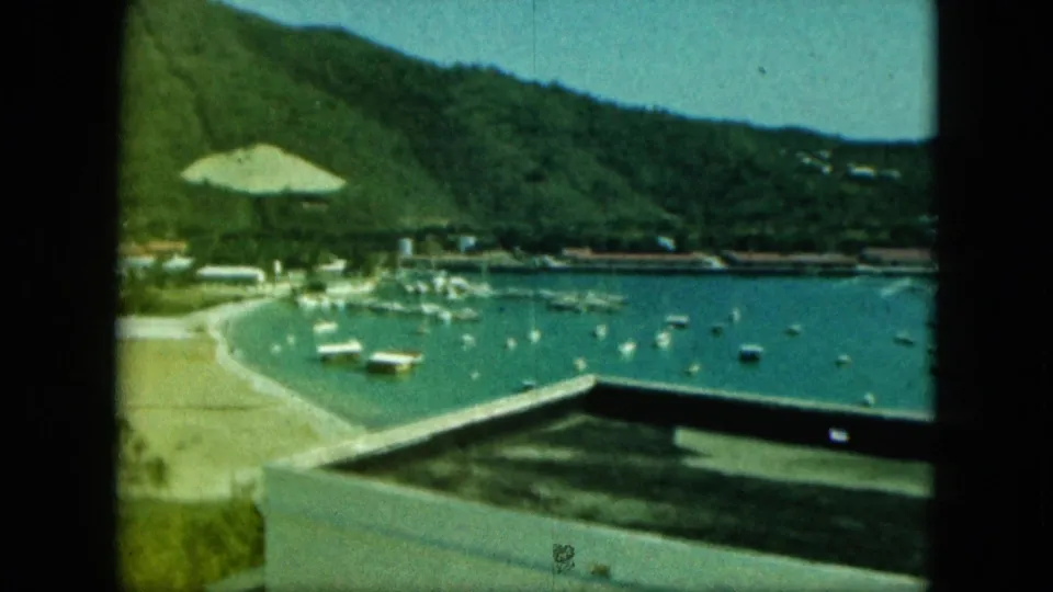 1964 8mm harbor view with turquoise water, small anchored boats, a beach edge, green hills, and foreground structure.