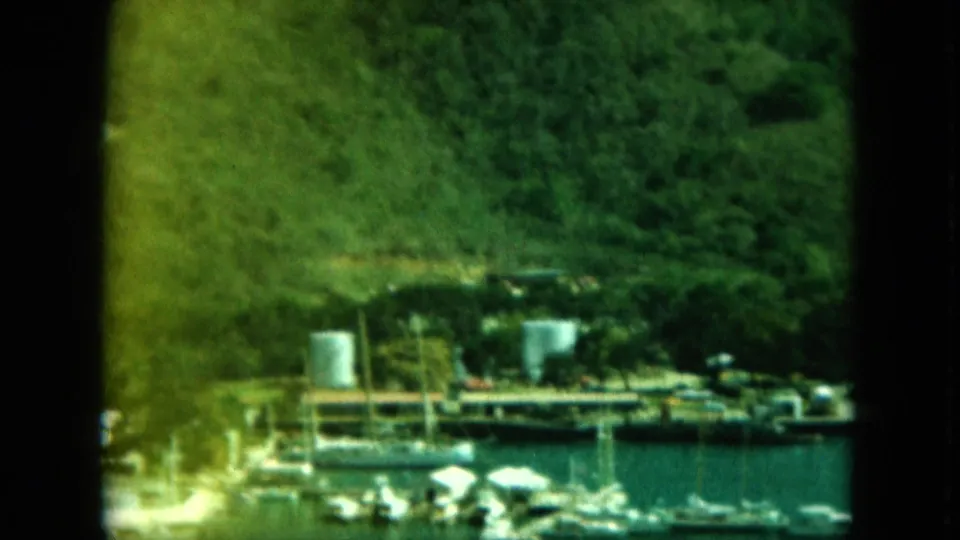 Distant harbor detail showing boats, dock structures, cylindrical tanks, shoreline activity, and steep green hills in 1964 8mm footage.