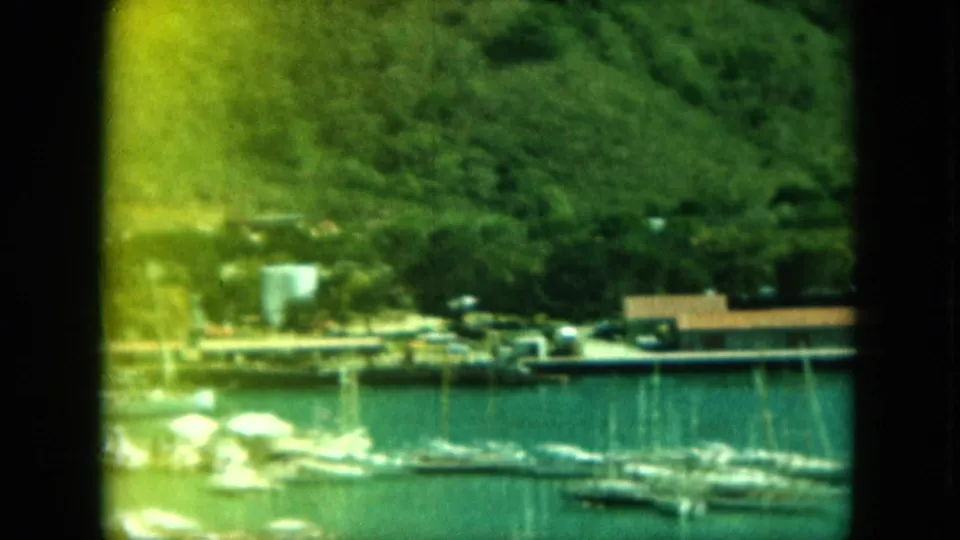 1964 8mm home-movie frame of a Saint Thomas harbor with sailboats near docks below a green hillside and low waterfront buildings.