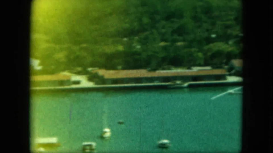 Final 1964 8mm frame of a Saint Thomas harbor pan, with small boats on open water and low waterfront structures under green hills.