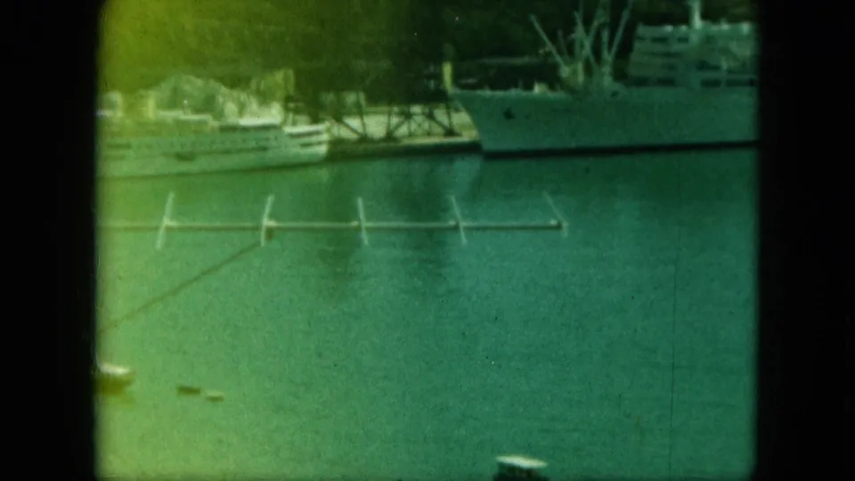 Faded 8mm frame of a large vessel and smaller boats across calm harbor water, with docks and shoreline in the background.