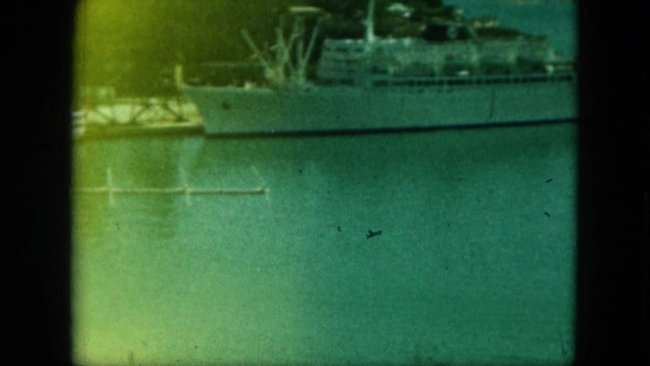 Docked ship viewed across calm water with small moored boats nearby.