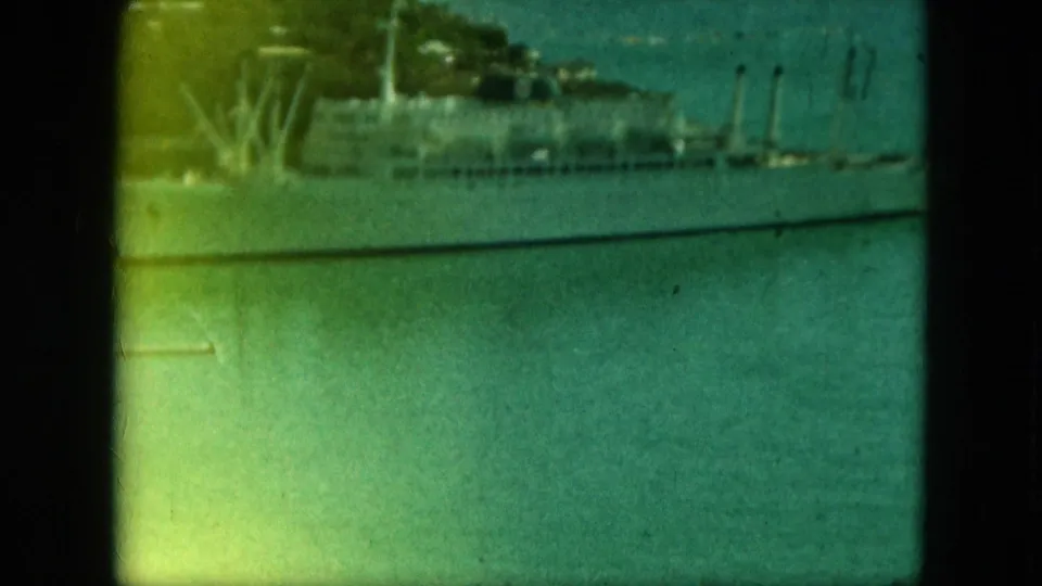 Soft, faded 8mm frame showing part of a large docked vessel, narrow docks, water, and trees along the harbor edge.