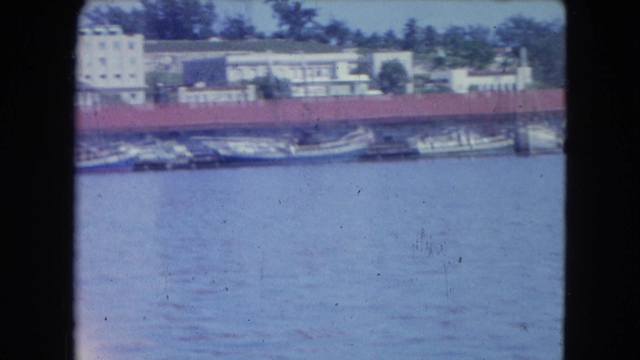 Docked boats across blue harbor water below pale buildings and a long red-roof structure.