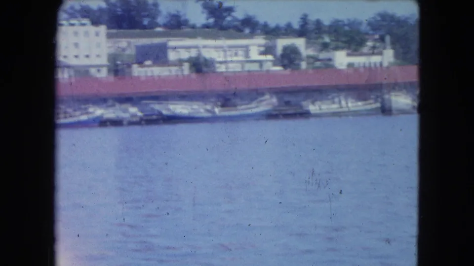1964 San Juan waterfront view with small boats moored beside red-roof industrial buildings across calm harbor water.