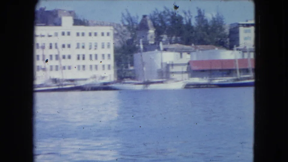 San Juan harbor frame with a white multi-story waterfront building, moored boats, trees, and a red-roof dock structure.