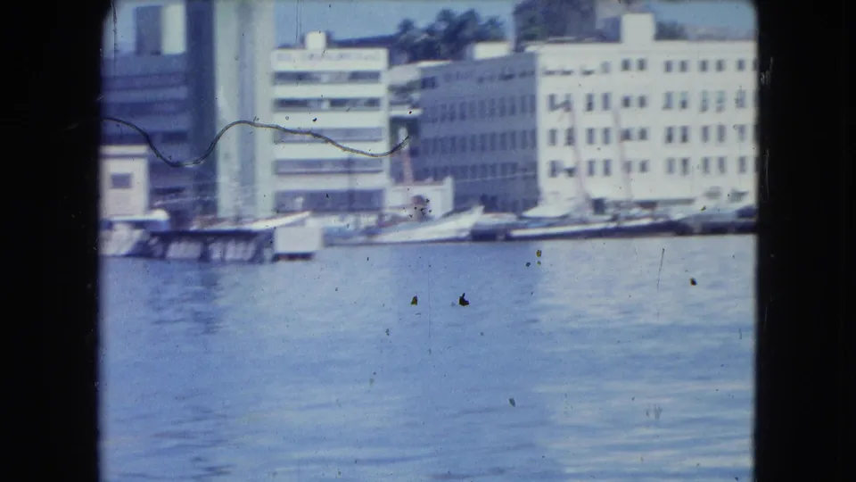 1964 San Juan waterfront view with pale industrial buildings, small boats, and rippled harbor water in a scratched 8mm frame.