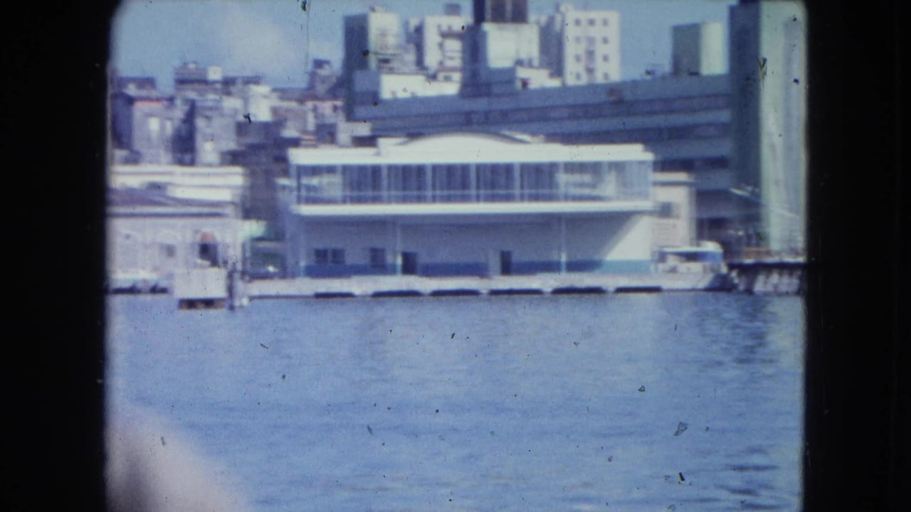 San Juan harbor view from a moving boat, showing a white waterfront building, pier line, water and mid-century city blocks.