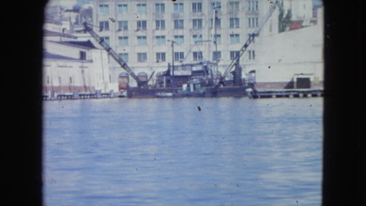 Waterfront buildings with a crane and barge across calm harbor water.
