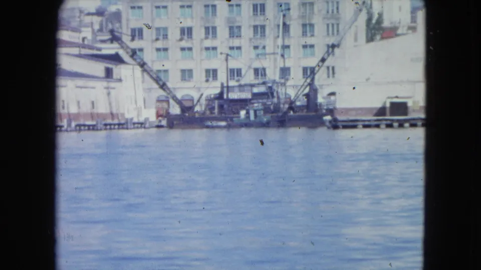 1964 San Juan harbor frame showing a large crane, docked barge, and waterfront buildings across blue water.
