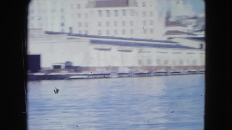 Soft 8mm frame of San Juan's inner harbor, with a long waterfront wall, pale buildings, and blue water.