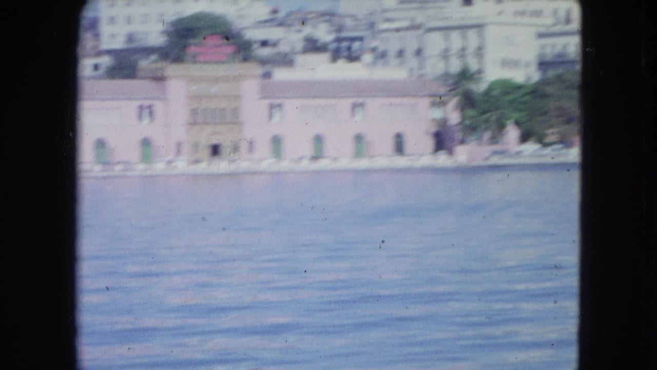 1964 San Juan harbor view from the water, with a pink waterfront building, green openings, palms, and distant city structures.