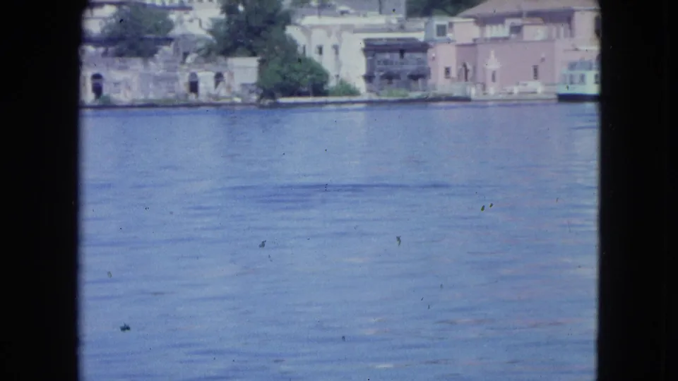 1964 waterfront pass-by in San Juan, showing rippled harbor water, pale shoreline buildings, trees, and a darker pier or balcony structure.