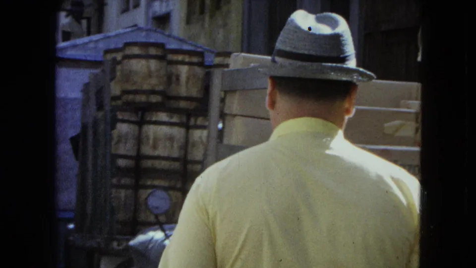 1964 San Juan street view of a man seen from behind in a light shirt and fedora walking past stacked barrels or boxes.