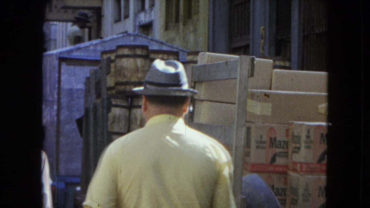 Man in yellow shirt and fedora walking past stacked boxes on a narrow street.