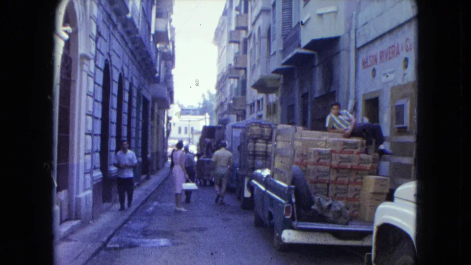 Wide 1964 San Juan street scene with pedestrians, a pickup loaded with cartons, and a person seated on the rear of the vehicle.