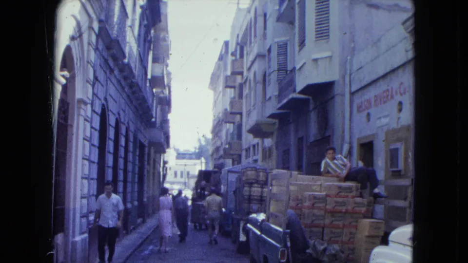 San Juan street view with pedestrians passing a pickup truck loaded with cartons and a man resting on the rear of the vehicle.