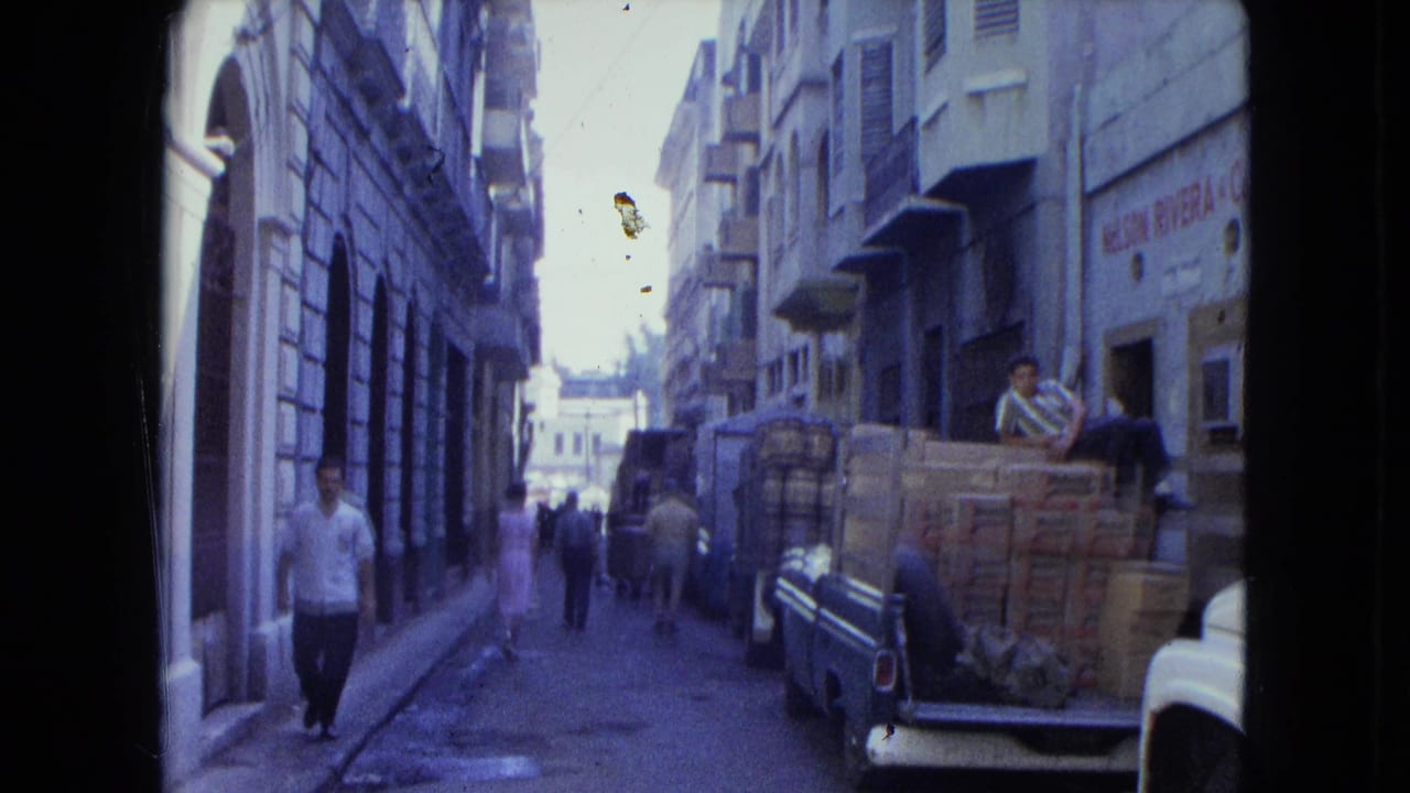 1964 San Juan street view with a man in a white shirt walking toward the camera near pickup trucks loaded with boxes.