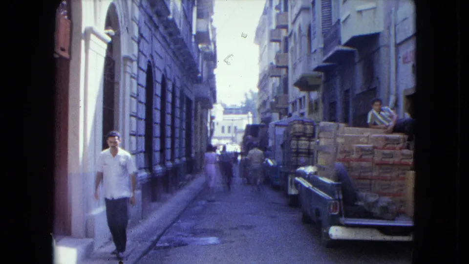 In San Juan, a man in a white shirt walks along a sunlit lane beside loaded pickup trucks and masonry buildings.