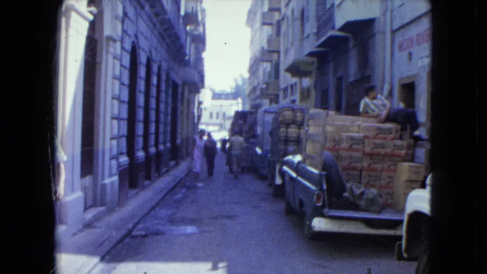A narrow San Juan street shows pedestrians moving past parked pickup trucks carrying stacked boxes in a commercial area.
